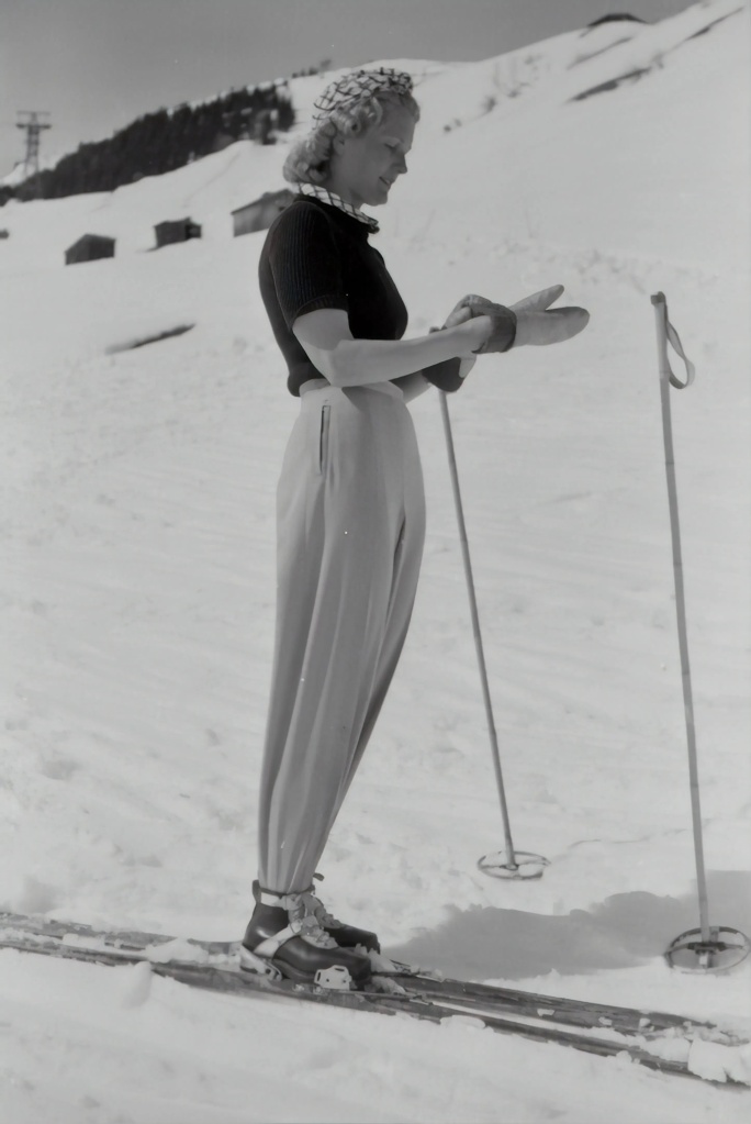 woman standing on skis of pre-1940 vintage, in light ski pants and short-sleeve top, with a snow-covered mountain slope behind